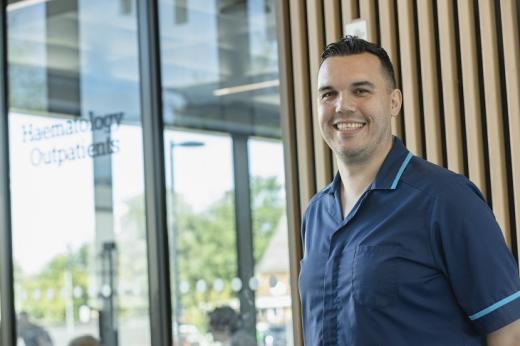 A man in a blue nurse outfit smiling in front of the modern Oak Cancer Centre building with wood panelling and glass