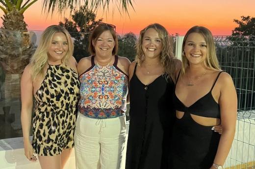 Four women on holiday, standing together under a pretty orange sunset with palm trees in the background