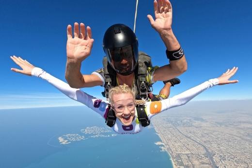 Two people, including an instructor with a helmet on and a fundraiser wearing a Royal Marsden Cancer Charity shirt, skydiving through mid air, with incredible views of the coast