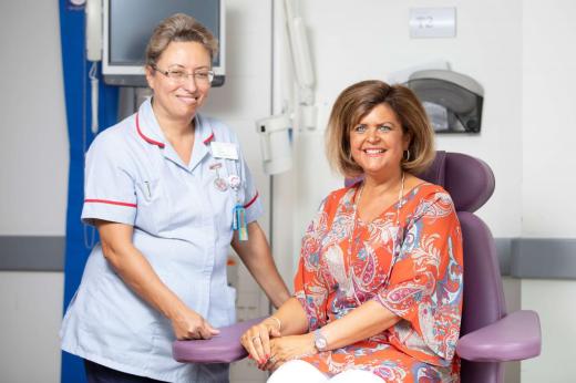 Louise and Senior Nurse Pilar in The West Wing. Louise is sat next to the Nurse in a purple treatment chair. She is wearing a bright orange and pink patterned shirt and both are smiling. 