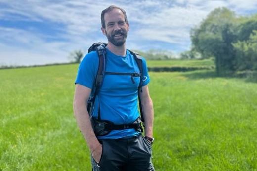 Jon standing in a rolling green field on a sunny day, wearing a backpack and walking gear.