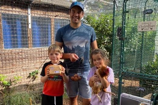 A man with his two young children standing in a chicken pen holding eggs, one child is holding a chicken