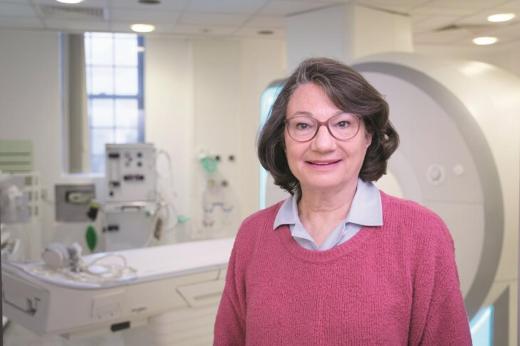 A Royal Marsden patient standing in front of an MRI machine in a hospital room, wearing a bright pink jumper and pink glasses