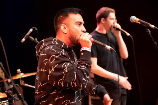 A young person performing into a microphone with a drum kit and another person singing behind them