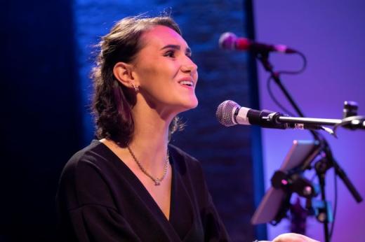 A young person dressed formally on a stage smiling and singing into a microphone
