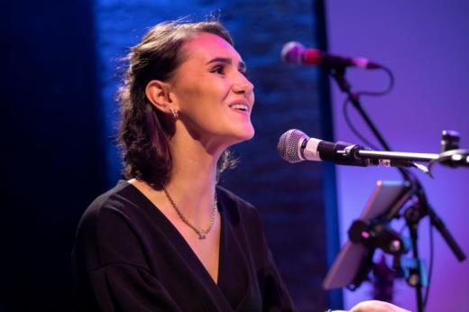 A young person dressed formally on a stage smiling and singing into a microphone 