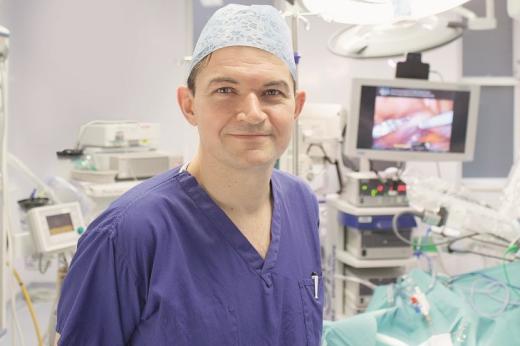 A Consultant Surgeon in medical scrubs, smiling and standing in an operating room in front of surgical equipment