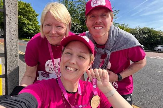 A Royal Marsden patient in a wheelchair smiling and holding up a Banham Marsden March medal, with two people smiling behind her.