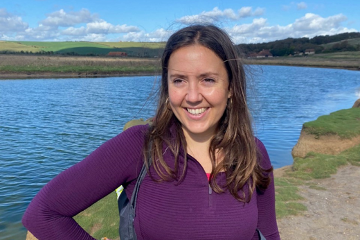 Tabitha smiling, wearing purple hoodie and standing in front of a lake