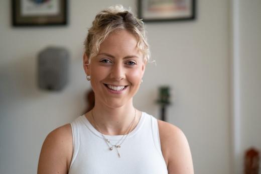 The head and shoulders of a young woman smiling, wearing her hair tied up and a white summer top with gold jewellery