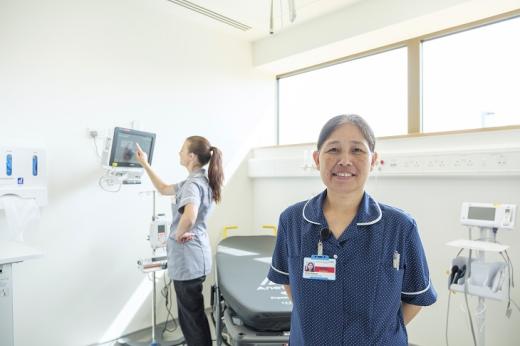 A nurse wearing a blue uniform and name badge smiling and standing in a bright, airy medical room in the Oak Cancer Centre. With a another nurse working in the background.