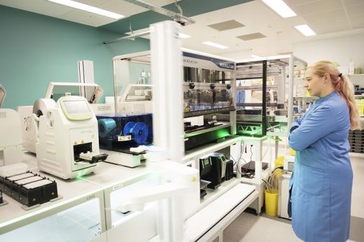 A large white robot with a moving arm picking up slides, in a Royal Marsden laboratory. It is being watched by a member of the lab team.
