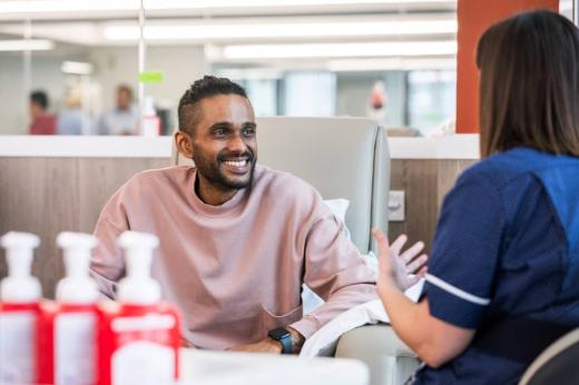 A man in a pink sweatshirt sat in a hospital chemotherapy bay. He is smiling and speaking with a nurse