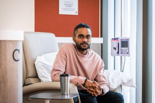 A man with short dark hair and a pink sweater sitting in a chemotherapy treatment chair in a hospital room.