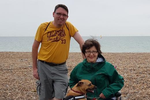 A man and a woman with a dog in front of a beach. They are both smiling, the woman is in a wheelchair and the dog has its legs on her lap