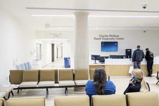 A pristine, white and airy waiting room with a front desk. There are some patients sitting on leather chairs and a couple talking to someone behind the front desk.