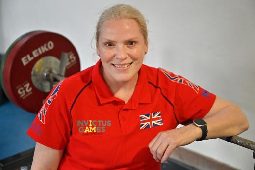 Amanda smiling and wearing a bright red Invictus Games UK shirt. She's leaning on a large weight in a gym.