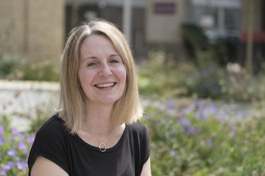 A Royal Marsden patient sitting in the flowery front garden of the Oak Cancer Centre. She has short blonde hair and a black top, she's smiling.
