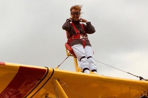 Marilyn on the wing of the plane, she is strapped in with a safety harness and wearing goggles. Her hair is blowing in the wind