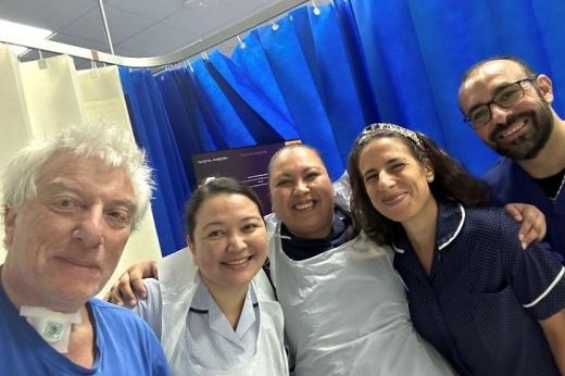 Mike, with a tracheostomy tube in his neck standing with a group of smiling staff at The Royal Marsden. They are all wearing scrubs and standing in a hospital room.