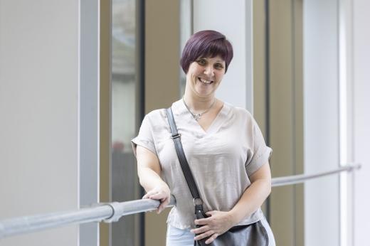 Roxanne, bowel cancer patient at The Royal Marsden, standing and smiling in front of the bright window of the Oak Cancer Centre.. She has short, purple/brown coloured hair, and is wearing a light top with a black handbag over her shoulder.