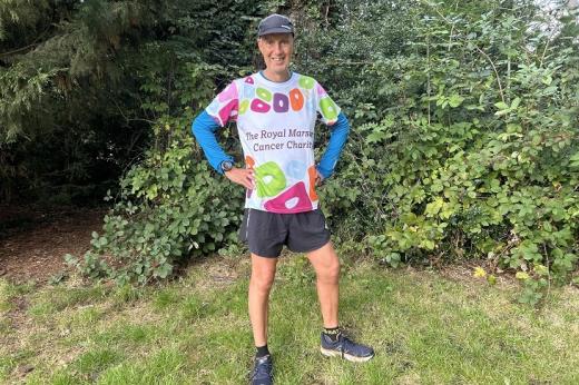 Kevin standing on a lawn with bushes and trees behind him. He's wearing running shoes, shorts and a Royal Marsden Cancer Charity-branded shirt. 