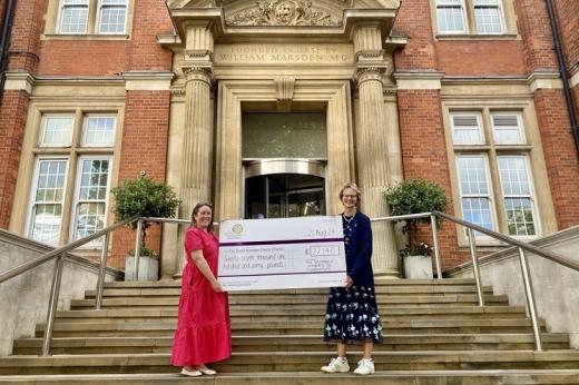 Two people standing on front steps of The Royal Marsden in Chelsea. They are wearing summery, flowery dresses and holding a large cheque for £27,140, dedicated to The Royal Marsden Cancer Charity.