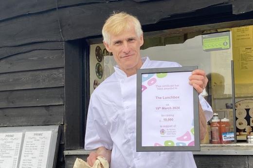 A man in a white chef shirt. He is holding up a Royal Marsden Cancer Charity certificate for raising £10,000. He is standing outside a window hatch of a small lunch outlet.
