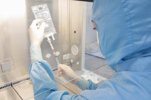 A Royal Marsden Nurse working in a sterile cellular therapies laboratory. She is wearing a blue sterile suit, gloves and mask and is using a syringe to pull liquid from a medication bag.