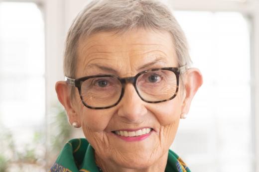 Head and shoulders of a woman with short grey hair, she's smiling and wearing stylish glasses and dark blue and green scarf