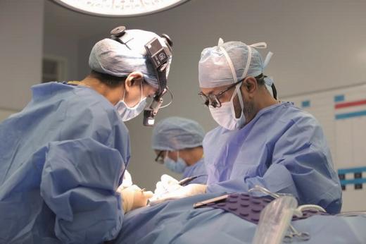Professor Paleri looking down and conducting surgery on a patient. He is opposite another surgeon, they both have masks, hairnets and lights on their head