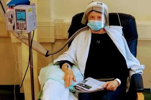A woman sat in a treatment chair holding a magazine. She has a towel around her shoulders and is wearing a blue medical mask