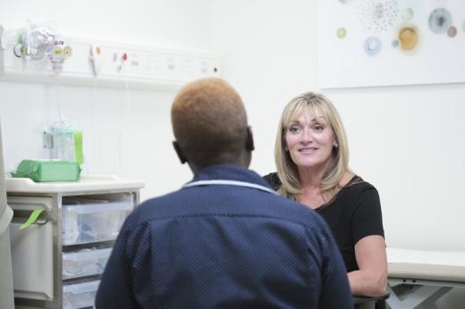 Two women talking. One is in a nurse's uniform and has her back to us, the other is wearing a black top and is facing forwards