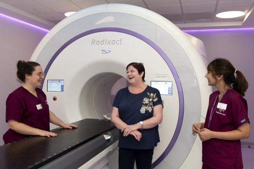 A patient smiling and laughing with two people from The Royal Marsden's radiotherapy team. They are standing in front of a large white Radixact machine, which has a lateral bed for patients to lie down and a tunnel, where patients' heads go.