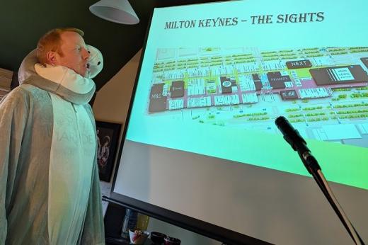 Tom wearing an animal onesie and talking into a mic about a PowerPoint slide named 'Milton Keynes - the ISghts'