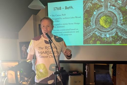 Tom in a Royal Marsden Cancer Charity-branded t-shirt, talking to a camera. He is standing in front of a large projector, showing PowerPoint slides of information on 1786 Bath. 