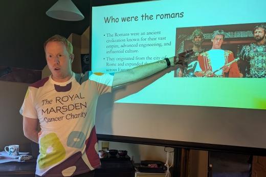 Tom in a Royal Marsden Cancer Charity-branded t-shirt, pointing to a large screen with a PowerPoint that says 'Who were the Romans'.