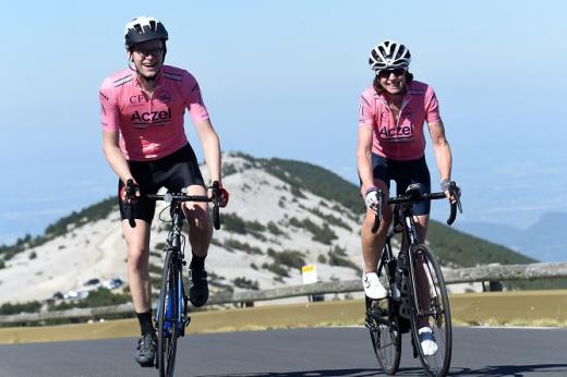 Two people in pink Le Cure branded cycling gear, cycling up a road-side mountain. It's a sunny day and there is a white-coloured mountain in the background behind them. 