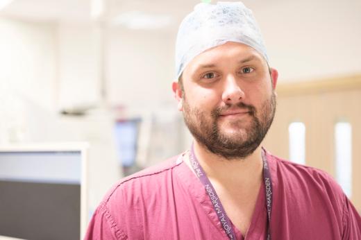 A man, Dr Ed Johnston, looks into the camera. he has a light brown beard and a blue scrub cap on. He is wearing pink scrubs and a Royal Marsden lanyard.