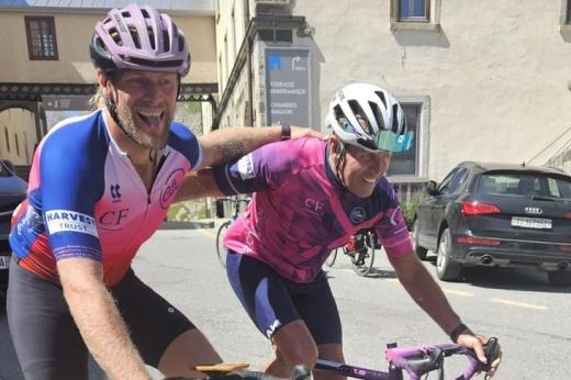 Two people, a father and a son, wearing cycling gear on bikes, they are smiling and have their arms around each other.