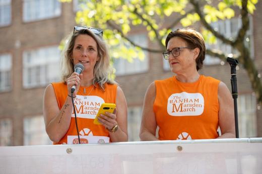 Two people with orange Banham Marsden March t-shirts standing on a podium at the start of the march at Dovehouse Green. One is speaking into a microphone, while holding a phone to refer to notes.