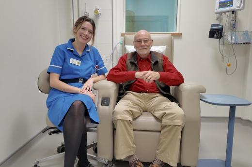 Patient, Richard, is smiling and sitting on a comfortable leather treatment chair in an empty, quiet medical room at The Royal Marsden. Sitting next to him in a swivel office chair is his nurse who is wearing a bright blue uniform and name badge.