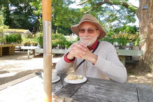 Richard sitting on a picnic table at a garden centre or plant nursery. It's a bright, sunny day and he's wearing a bucket hat with a shirt and light jumper, while enjoying a cup of coffee and one half of a buttered scone.