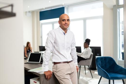 A man in white shirt and beige trousers, leaning against an office desk