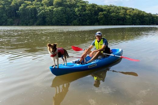 Dennis on a canoe, on a large river with trees in the background. He's wearing a bright yellow life jacket and his dog is standing on the front of the boat.