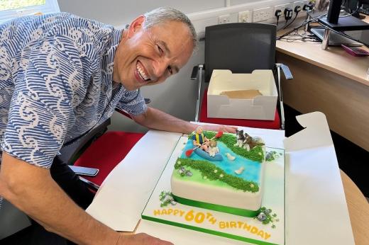 Dennis wearing a blue, pattered shirt. He's smiling and leaning over a 60th birthday cake that is decorated with icing figures of him and his dog canoeing down a river.