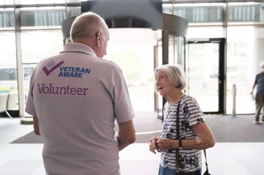 Brain in a light pink polar neck shirt that reads 'Veteran aware volunteer' on the back. He's speaking to a patient in the entrance of The Royal Marsden's Oak Cancer Centre