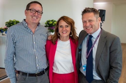 Shiona at The Royal Marsden, she's wearing a bright pink suit, white top and large hooped earrings. Shiona's standing in the middle of her husband, who is wearing a smart chequered shirt, and Professor James Larkin who is wearing a formal grey suit.