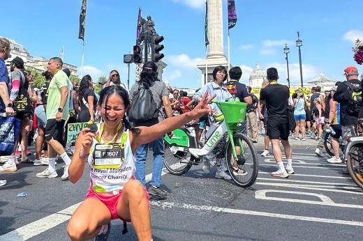 RMCC runner posing with her medal, crouched on the floor doing a peace sign to the camera with pedestrians in the background. 