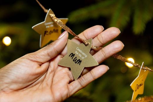 A close up photo of a hand holding a golden star decoration, hung on a christmas tree. The star says 'Celebrating the life of Mum' with the word 'mum' handwritten on the star.